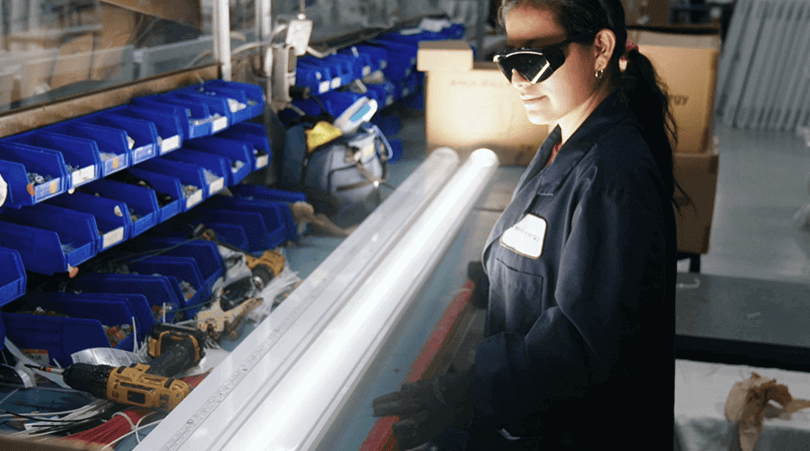 Female employee wearing blue lab coat testing a white linear LED light fixture in a manufacturing plant